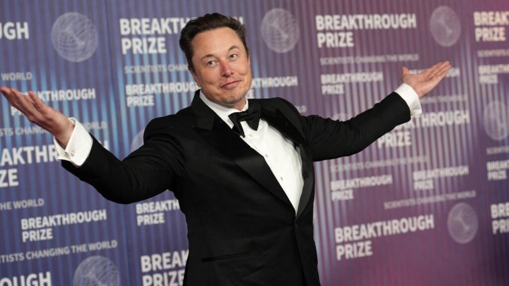 Elon Musk stands with open arms at the Breakthrough Prize event, wearing a classic black tuxedo with a bow tie